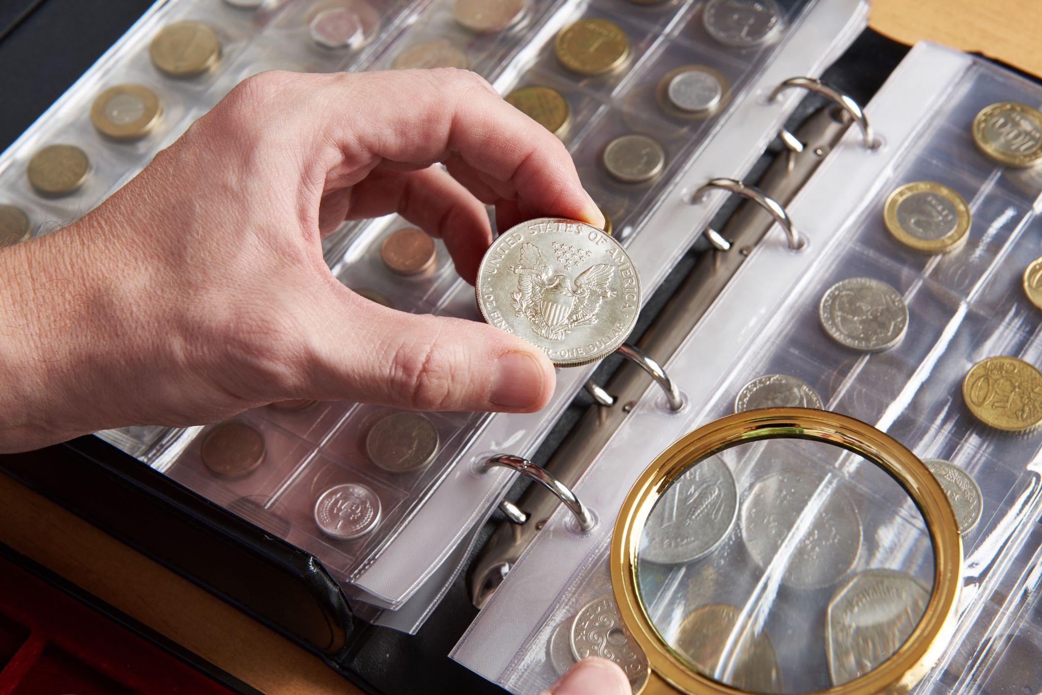 Man holding silver coin with left hand and binder with rare coins organized inside on table.